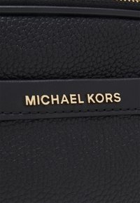 Bolso de cuero negro texturizado con el logo dorado "MICHAEL KORS", con acentos lisos y costuras visibles en los bordes.