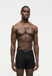 Μυώδης άνδρας με σκούρο δέρμα, όρθιος, φοράει μαύρα μποξέρ Calvin Klein, σε απλό φωτεινό γκρι φόντο.