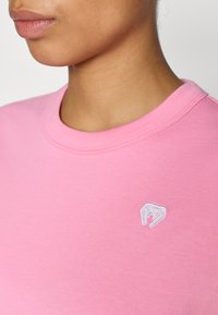 Ružové bavlnené tričko s okrúhlym výstrihom. Na ľavej strane logo vyšívané bielou niťou. Hladká textúra a priliehavý strih.