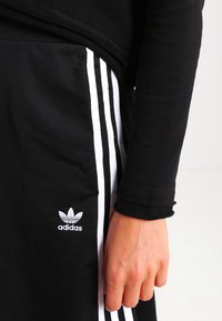 Crne sportske hlače s bijelim bočnim trakama, s izvezenim bijelim Adidas logotipom na lijevom bedru. Glatka tkanina, prilagođeni dizajn.