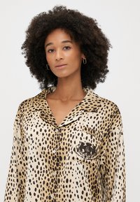 Top de pijama din satin cu mânecă scurtă, cu imprimeu animalier bej și negru, având un buzunar aplicat cu un logo și nasturi rotunzi pe față.