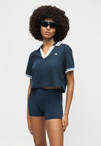 Γυναίκα με μεγάλο σγουρό μαλλί, φοράει κοντομάνικη μπλε σκούρα πόλο μπλούζα με λευκή μπορντούρα, μπλε σκούρα σορτς με ψηλή μέση και σκουρόχρωμα γυαλιά ηλίου τύπου wraparound, όρθια.