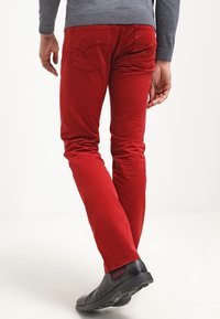 Pantaloni rossi in cotone con vestibilità slim, dotati di tasche posteriori e una texture liscia. Indossati con scarpe nere senza lacci.