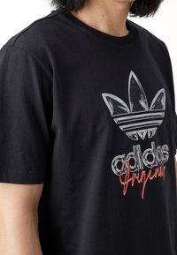 Černé bavlněné tričko s velkým stříbrným a bílým logem Adidas ve vintage designu a červeným textem "Originals" pod ním.