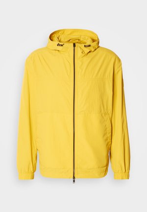 Κίτρινο μπουφάν αδιάβροχο με φερμουάρ, ρυθμιζόμενο κουκούλι και ελαστικά μανίκια, κατασκευασμένο από ελαφρύ, αδιάβροχο υλικό.