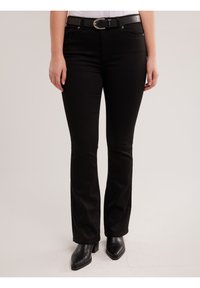 Sieviete valkā melnas piegulošas džinsas, melnu jostu ar sudraba sprādzi un melnas papēžainas potīšu zābakus, stāvot pret vienkāršu fonu.