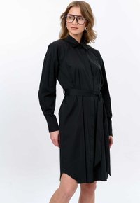 Schwarzes Hemdkleid mit langen Ärmeln, knöpfbarem Vorderteil und tailliertem Gürtel. Mit seitlichen Schlitzen und weichem, glattem Stoff.