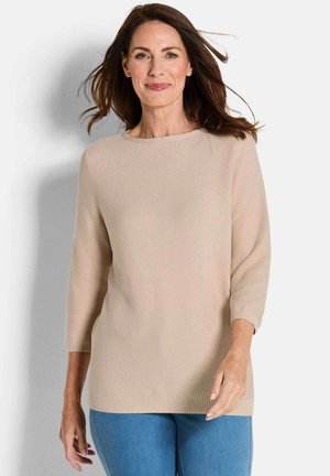Pull en tricot beige avec des manches trois-quarts, un col rond et un ourlet côtelé. Texture douce, coupe décontractée. Porté par-dessus un jean bleu clair.