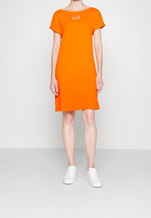 Personne debout portant une robe orange vif à manches courtes, longueur aux genoux, et des baskets blanches sur un fond clair uni.