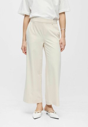 Žena oblečená v béžových širokých nohaviciach s vysokým pásom, bielom krátkym tričku, bielych nízkych topánkach s špicatou špičkou a strieborným náramkom na pravej ruke.