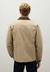 Giacca beige con colletto in corduroy marrone, maniche lunghe e taglio dritto. Presenta una cucitura semplice e polsini con bottoni per un tocco di dettaglio.