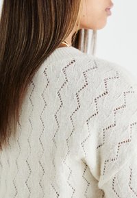 Maglione bianco lavorato a maglia con motivo a zig zag e dettagli in pizzo, scollo a coste e vestibilità rilassata. Tessuto morbido e texturizzato.