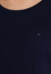 Marinblå bomullströja med rund halsringning, som har en liten broderad logotyp med röda, vita och blå detaljer på bröstet.