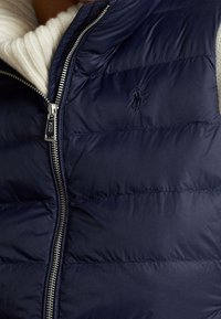 Nära bild av en marinblå puffväst med en silverfärgad dragkedja, lagd över en vit, ribbad stickad tröja. Polo-logotypen synlig på västen.