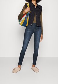 Svart quiltad väst, olivfärgad långärmad tröja, mörkblå skinny jeans, beige tofflor med detaljer. Färgglad vävd handväska med ränder.