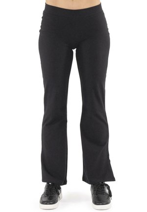 Une personne porte un pantalon noir évasé et des baskets noires à semelles blanches, et se tient devant un fond blanc uni.