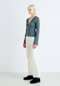 Långärmad, figurnära topp i skifferblått, i omslag-stil, kombinerad med krämfärgade utsvängda byxor och svarta slip-on skor, buren av en modell mot en ljus bakgrund.