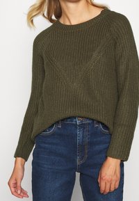 Olivengrüner Strickpullover mit einem strukturierten Rippenmuster, Rundhalsausschnitt und langen Ärmeln, kombiniert mit dunkelblauen Jeans.