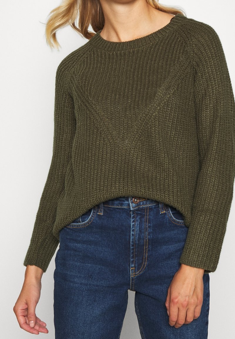 Olivengrüner Strickpullover mit einem strukturierten Rippenmuster, Rundhalsausschnitt und langen Ärmeln, kombiniert mit dunkelblauen Jeans.