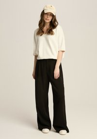 Biały sweter dzianinowy z krótkim rękawem, czarne spodnie z szerokimi nogawkami i beżowa czapka z haftowanymi detalami. Całość dopełniają casualowe buty.