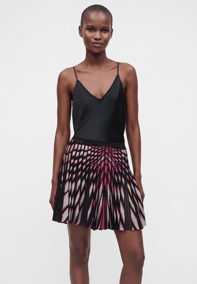 Femme portant un haut noir sans manches et une jupe plissée avec un motif géométrique noir, rose et blanc sur fond uni.