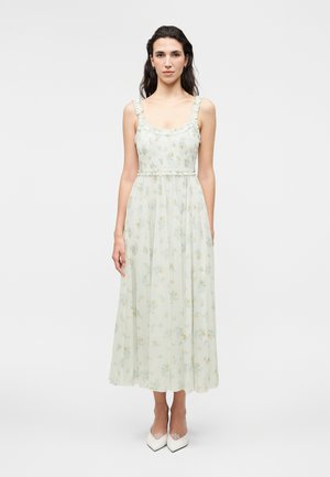 Nő áll, fehér virágos ujjatlan maxi ruhát visel fodros pántokkal és fehér hegyes orrú cipővel egyszerű háttér előtt.