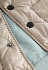 Veste matelassée beige avec boutons-pression noirs, portée par-dessus un tissu texturé vert clair. Présente une surface en nylon lisse et des sections de couleur contrastante.