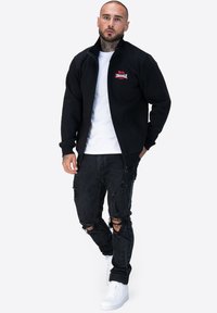 Giacca nera con zip in pile con un logo rosso sul petto, abbinata a jeans neri strappati e sneakers bianche. Design semplice, vestibilità casual.