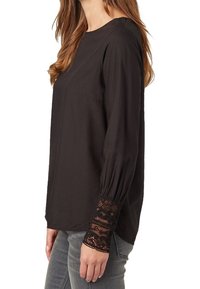 Schwarze Bluse mit langen Ärmeln und Rundhalsausschnitt, mit strukturierten Spitzenakzenten an den Bündchen und einem lässigen Schnitt, kombiniert mit grauen Jeans.