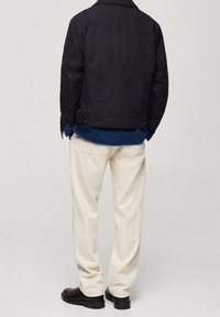 Granatowa kurtka z kołnierzem, noszona na niebieskiej koszuli, w połączeniu z kremowymi spodniami i czarnymi butami. Minimalistyczny design, jednolite kolory, swobodny krój.