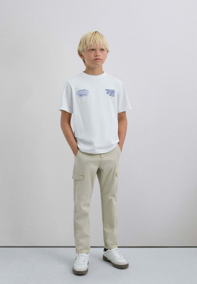 Weißes Baumwoll-T-Shirt mit blauen Druckgrafiken, kombiniert mit beigen Cargo-Hosen mit Seitentaschen. Weiße Sneaker mit grauen Akzenten.