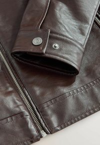 Manche d'une veste en cuir marron foncé avec boutons-pression argentés et une fermeture éclair argentée adjacente sur une surface en cuir texturée.