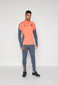 Atletiline treeningpükskostüüm, millel on erk korallivärvi särk hallide varrukatega ja sobivad hallid püksid. Komplektis on kolm triipu ja logo detailid.