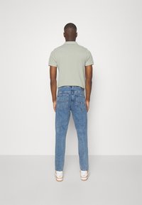 Světlé zelené polo tričko s límečkem, v kombinaci se světle modrými džínami a bílými teniskami, pohled zezadu na jednobarevném pozadí.