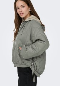 Tikkitakki oliivinvihreänä, vaaleanbeige huppu, säädettävä kiristysnauha helmassa ja sivutaskut. Pehmeä, tasainen pinta.