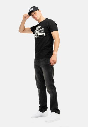 Zwarte katoenen T-shirt met wit logo, gecombineerd met donkere jeans en witte sneakers, aangevuld met een zwarte pet met logo-detail.