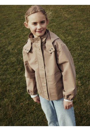 Junges Mädchen steht auf dem Gras, trägt eine beige Kapuzenjacke und hellblaue Jeans, schaut leicht zur Seite mit einem sanften Lächeln.