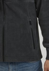 Fleecejacke in Dunkelgrau mit glatter Textur, ausgestattet mit einer seitlichen Reißverschlusstasche und gerippten Bündchen. Das Design umfasst flache Nähte.