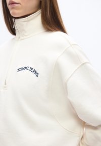 Γυναίκα που φοράει εκρού φούτερ με ψηλό γιακά και το κείμενο "TOMMY JEANS" κεντημένο με μπλε στο στήθος, κομμένο στο ύψος του στόματος και του ώμου.