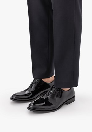 Zapatos de vestir de cuero negro pulido usados con pantalones negros a medida, mostrados desde la mitad de la pantorrilla hacia abajo sobre un fondo blanco.