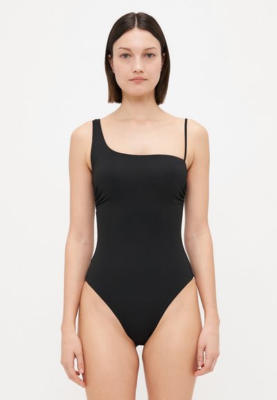 Femeie îmbrăcată într-un costum de baie negru dintr-o singură piesă cu bretele asimetrice, stând în fața unui fundal alb simplu.
