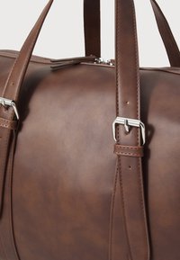 Bolso de viaje de cuero marrón con una textura suave, que cuenta con herrajes plateados y un diseño de doble correa para mayor comodidad al llevarlo.