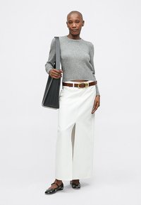 Suéter cinza em tricô, saia maxi de denim branca com uma fenda na frente, cinto castanho com fivela dourada e sapatos pretos slip-on. Mala de ombro preta.