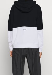 Nike-hoodie med en tvåfärgad design i svart och vitt, mjukt material och en broderad logotyp på baksidan, i kombination med mörka byxor.