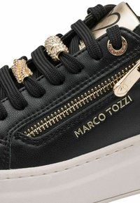 Baskets noires avec une semelle blanche épaisse, une fermeture éclair dorée et des perles dorées décoratives sur les lacets noirs, marquée "Marco Tozzi" sur le côté.