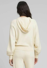 Suéter con capucha color crema, de corte relajado, con puños y dobladillo acanalados. Textura de punto suave, visto desde atrás, combinado con pantalones a juego.