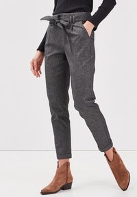 Kvinna som bär högmidjade grå paperbag-byxor med ett knytbälte, svart ribbat linne och bruna mocka ankelskor med låga klackar.