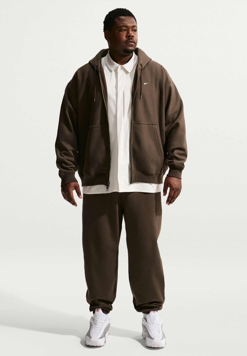 Hombre vistiendo una sudadera con capucha con cremallera de Nike de color marrón oscuro y pantalones deportivos a juego, camisa blanca con cuello y zapatillas blancas, de pie frente a un fondo liso.