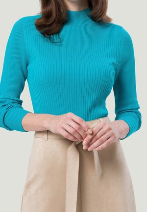Haut en tricot côtes turquoise avec col montant, manches longues et coupe ajustée, accompagné d'une jupe en suède beige à taille nouée.