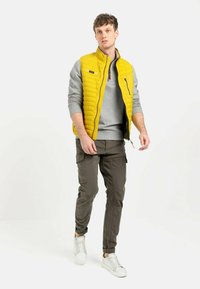 Gul quiltet vest med grå ærmer, grå sweatshirt under, olivengrøn cargo-bukser og hvide sneakers. Har en lynlåslomme på vesten.